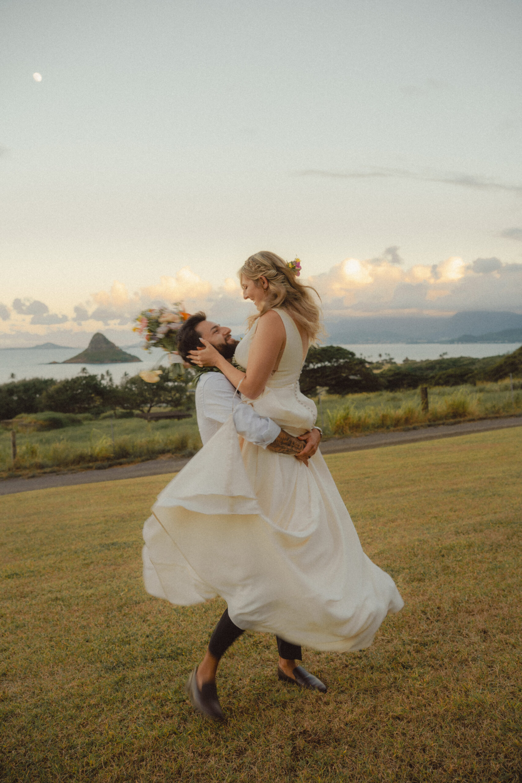 Bride and Groom dancing in a field during sunset with ocean backdrop at Kualoa Ranch.